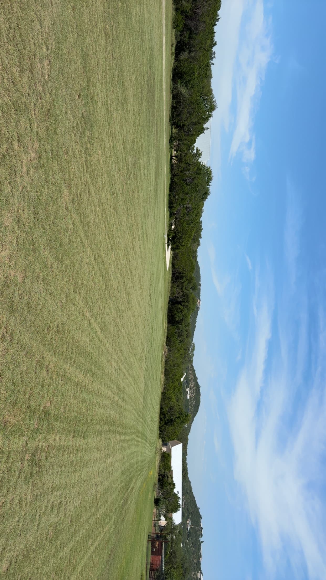 Expansive green field bordered by trees with distant hills under a blue, wispy sky.