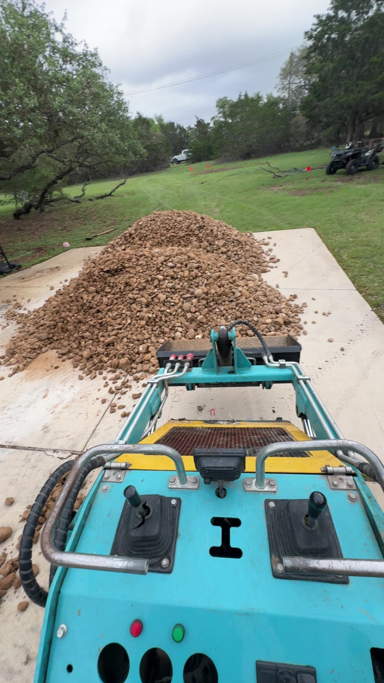 Teal loader controls facing a large pile of brown rocks on a concrete driveway.
