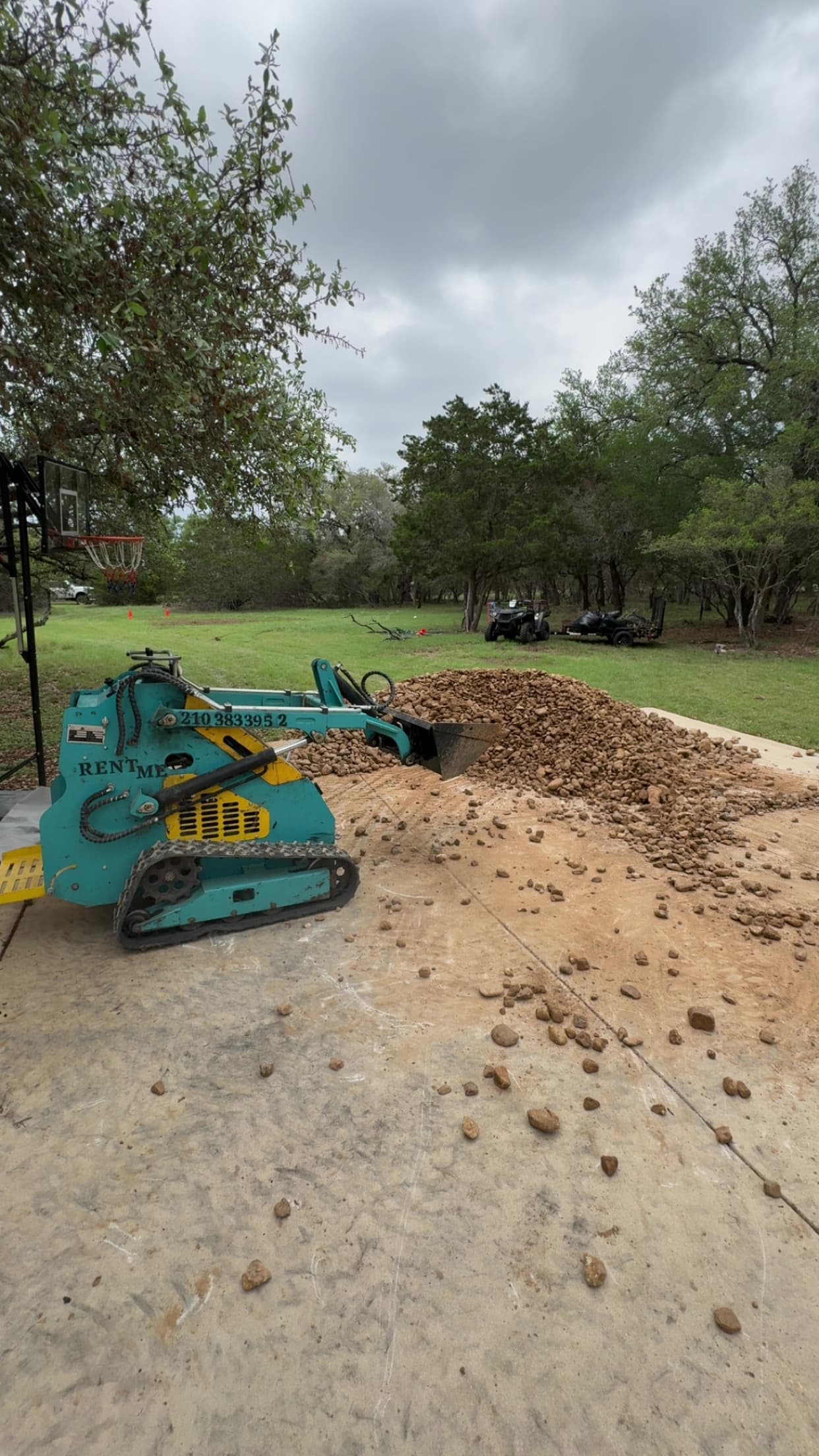 Teal mini skid steer moving a large pile of brown rocks on a concrete driveway.