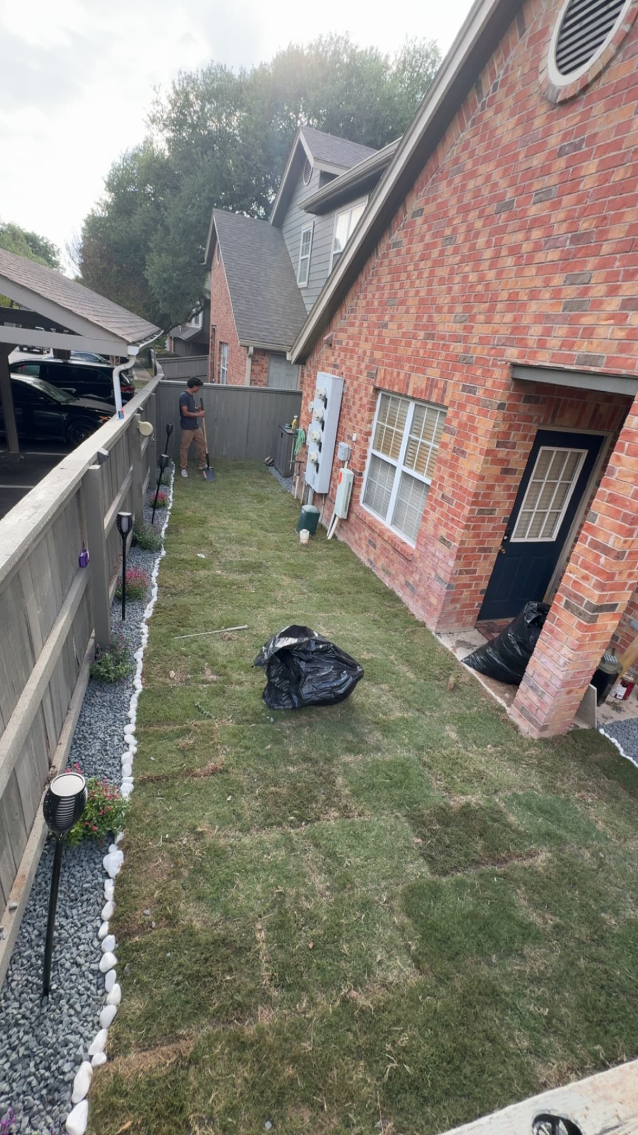 Man landscaping a narrow side yard with fresh sod beside a red brick house.