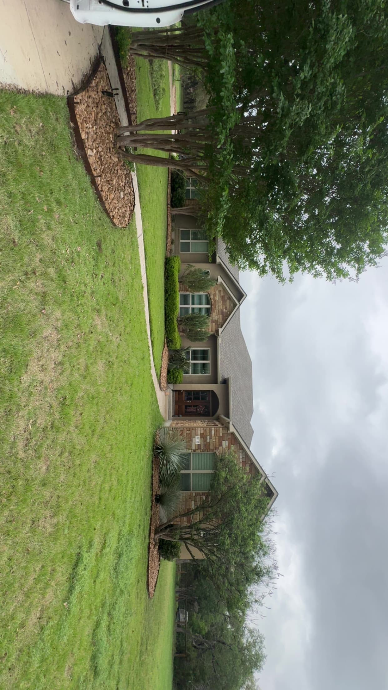 Suburban stone house with a green lawn, curved walkway, and trees on an overcast day.