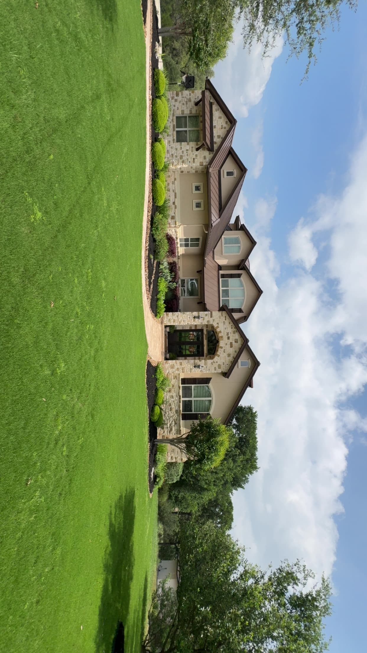 Large stone and stucco house with brown metal roof, manicured lawn, and bright blue sky.