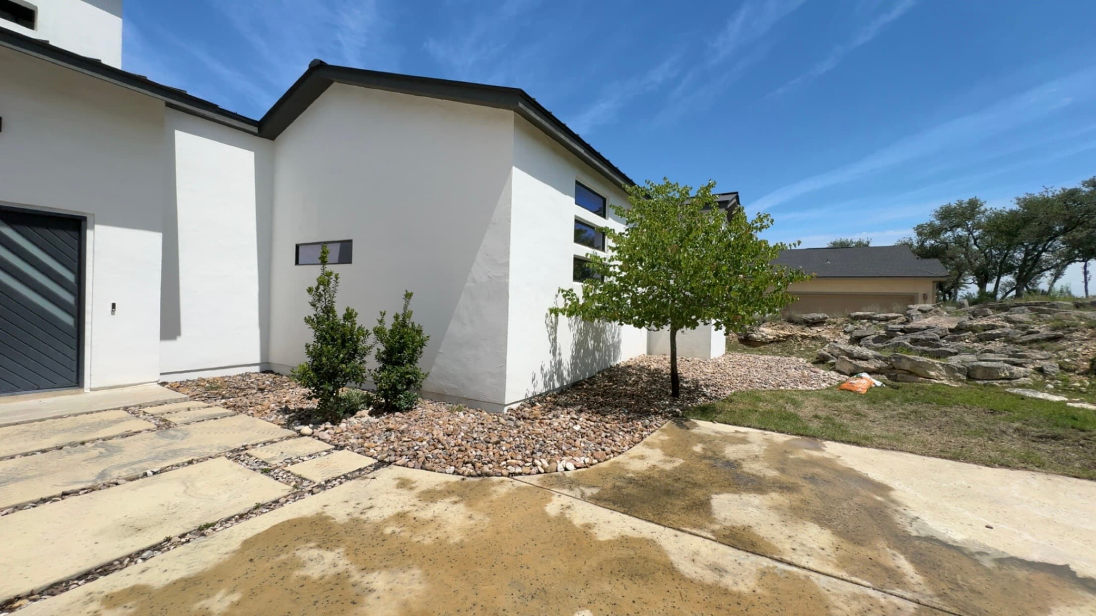 Modern white house with dark trim, small tree, and gravel landscaping under blue sky.